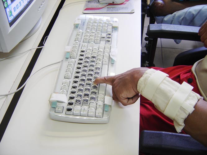 Foto com recursos de Tecnologia Assistiva: pulseira de pesos colocada no pulso de usurio, teclado com colmia e com letras destacadas com alto contraste.